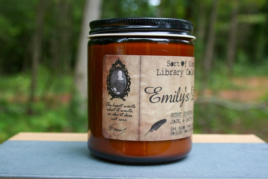 A white soy wax candle in an amber jar with a rectangular label against a white background. The label has like an aged paper look with a binding crease on the left side creating a column. The column has a black and white portrait of Dickinson in a black frame. Under the portrait is the quote "The heart wants what it wants, or else it does not care." followed by Dickinson's signature.