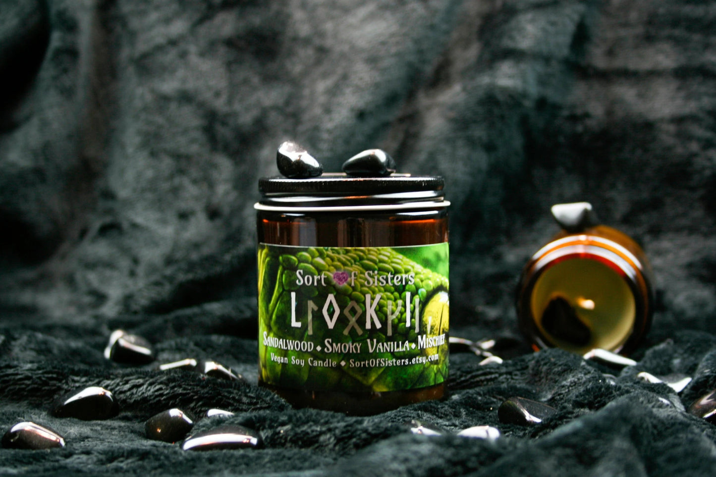 A green soy wax candle in an amber jar with a rectangular label. The label has a green snake on it with yellow eyes. White text on the label reads "Sort of Sisters" logo, below that reads "Loki" (in English and Ruins), below that reads "sandalwood, smoky vanilla, and mischief", and finally below that reads "vegan soy candle, sortofsisters.etsy.com". The candle is on a black velvet background surrouned by obsidian stones.