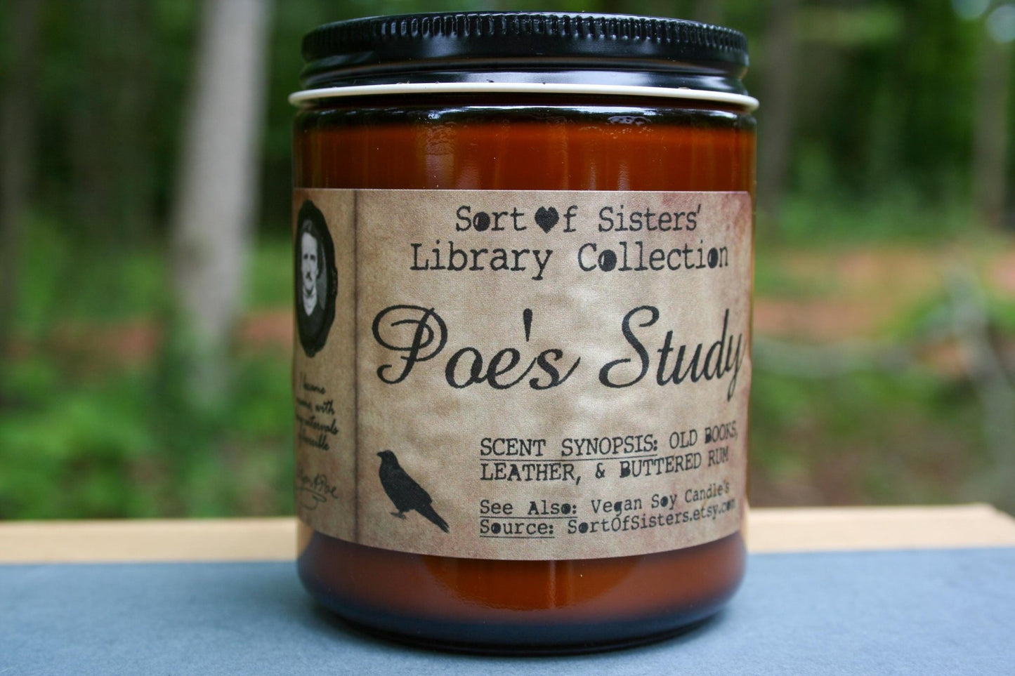 A white soy wax candle in an amber jar with a rectangular label against a white background. The label has like an aged paper look with a binding crease on the left side creating a column. The column has a black and white portrait of Poe in a black frame. Under the portrait is a quote and Poe's signature. In the larger right column text reads "Sort of Sisters" logo at the top, then "Poe's Study", then "old books, leather, & buttered rum", and below that "vegan soy candles, sortofsisters.etsy.com"