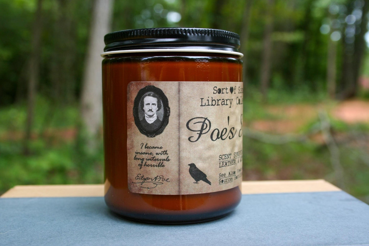 A white soy wax candle in an amber jar with a rectangular label against a white background. The label has like an aged paper look with a binding crease on the left side creating a column. The column has a black and white portrait of Poe in a black frame. Under the portrait is the quote "I became insane with long intervals of horrible sanity" followed by Poe's signature.