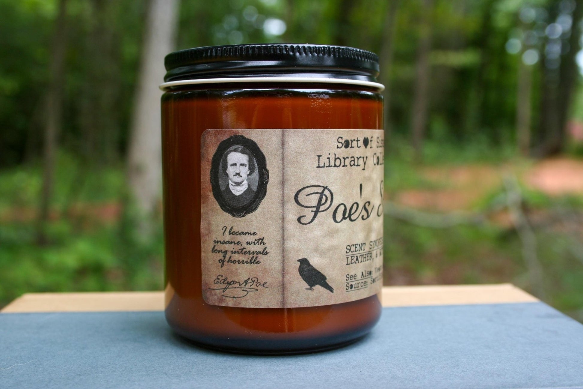 A white soy wax candle in an amber jar with a rectangular label against a white background. The label has like an aged paper look with a binding crease on the left side creating a column. The column has a black and white portrait of Poe in a black frame. Under the portrait is the quote "I became insane with long intervals of horrible sanity" followed by Poe's signature.