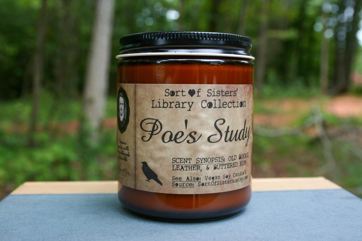A white soy wax candle in an amber jar with a rectangular label against a white background. The label has like an aged paper look with a binding crease on the left side creating a column. The column has a black and white portrait of Poe in a black frame. Under the portrait is a quote and Poe's signature. In the larger right column text reads "Sort of Sisters" logo at the top, then "Poe's Study", then "old books, leather, & buttered rum", and below that "vegan soy candles, sortofsisters.etsy.com"