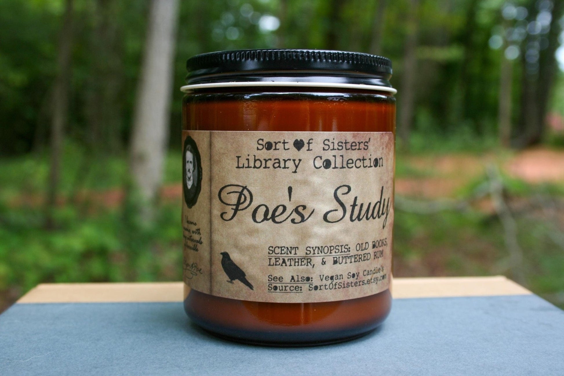 A white soy wax candle in an amber jar with a rectangular label against a white background. The label has like an aged paper look with a binding crease on the left side creating a column. The column has a black and white portrait of Poe in a black frame. Under the portrait is a quote and Poe's signature. In the larger right column text reads "Sort of Sisters" logo at the top, then "Poe's Study", then "old books, leather, & buttered rum", and below that "vegan soy candles, sortofsisters.etsy.com"