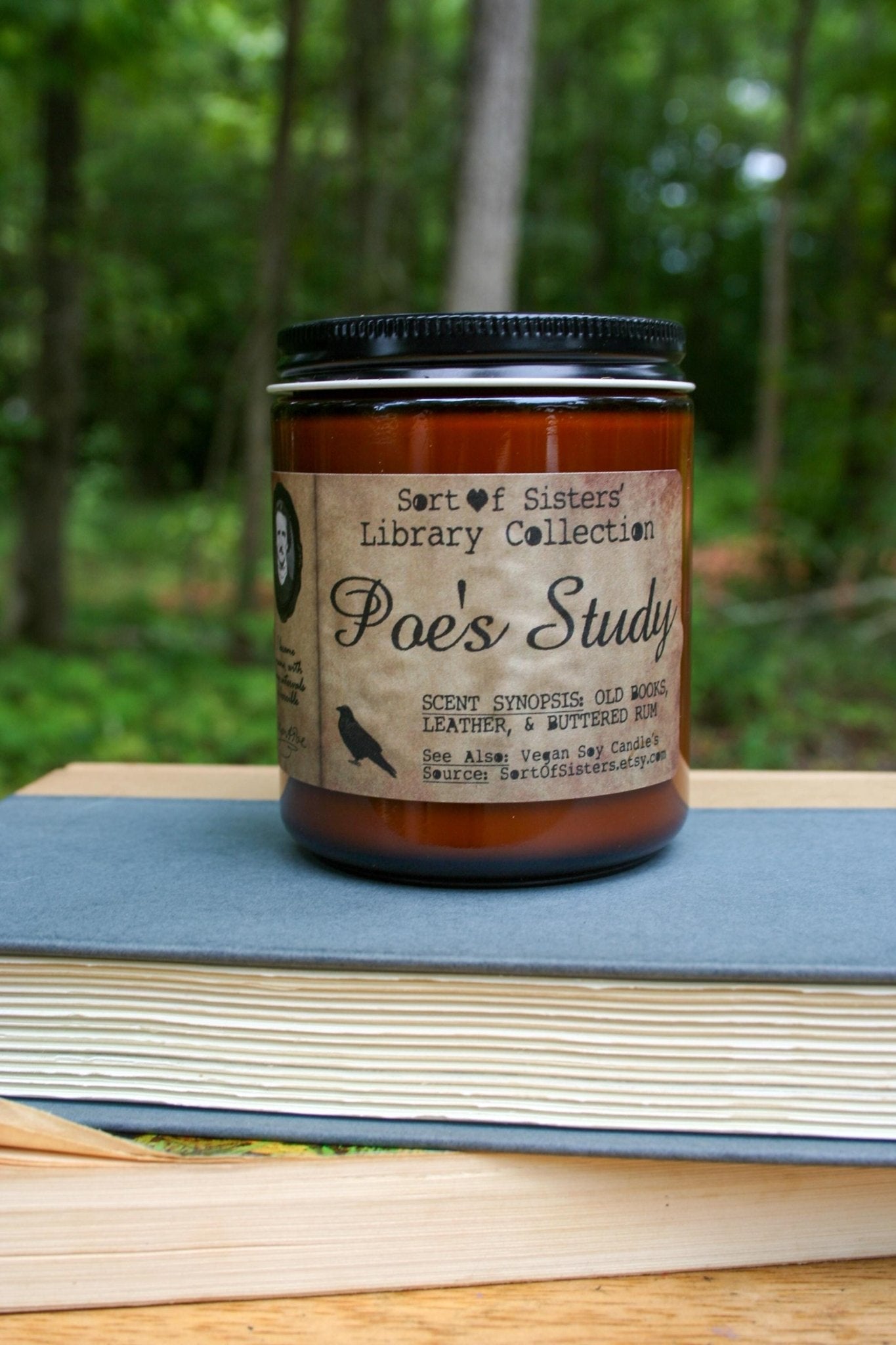 A white soy wax candle in an amber jar with a rectangular label against a white background. The label has like an aged paper look with a binding crease on the left side creating a column. The column has a black and white portrait of Poe in a black frame. Under the portrait is a quote and Poe's signature. In the larger right column text reads "Sort of Sisters" logo at the top, then "Poe's Study", then "old books, leather, & buttered rum", and below that "vegan soy candles, sortofsisters.etsy.com"