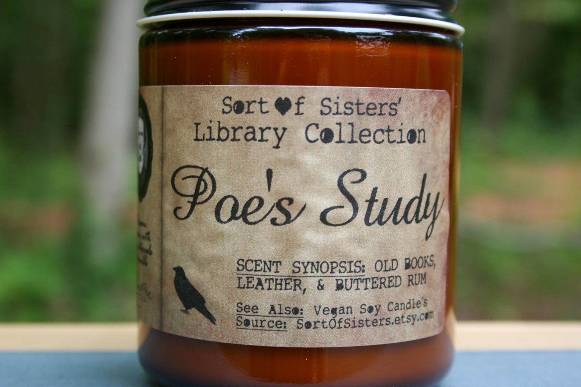A white soy wax candle in an amber jar with a rectangular label against a white background. The label has like an aged paper look with a binding crease on the left side creating a column. The column has a black and white portrait of Poe in a black frame. Under the portrait is a quote and Poe's signature. In the larger right column text reads "Sort of Sisters" logo at the top, then "Poe's Study", then "old books, leather, & buttered rum", and below that "vegan soy candles, sortofsisters.etsy.com"