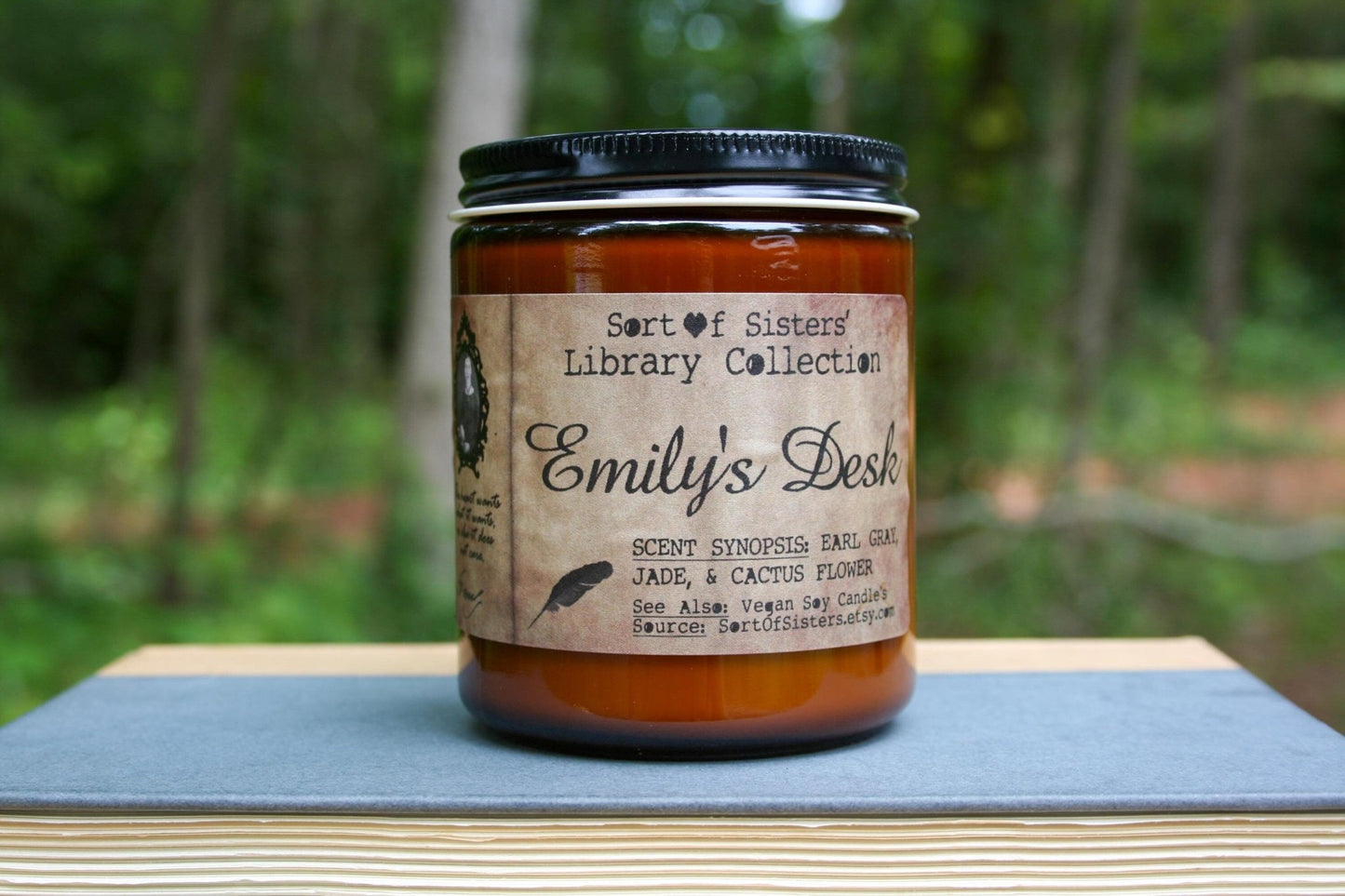 A white soy wax candle in an amber jar with a rectangular label against a white background. The label has like an aged paper look with a binding crease on the left side creating a column. The column has a black and white portrait of Dickinson in a black frame. Under the portrait is a quote and Dickinson's signature. In the larger right column text reads "Sort of Sisters" logo at the top, then "Emily's Desk", then "Earl Grey, Jade, & Cactus Flower", and below that "vegan soy candles, sortofsisters.etsy.com"