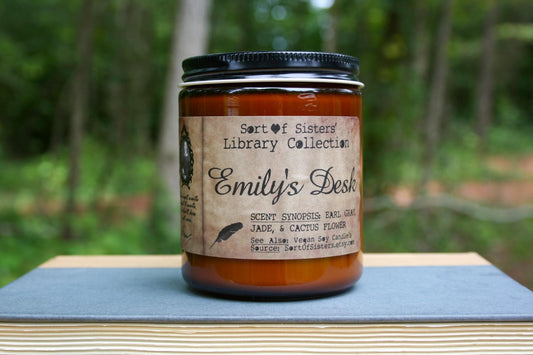 A white soy wax candle in an amber jar with a rectangular label against a white background. The label has like an aged paper look with a binding crease on the left side creating a column. The column has a black and white portrait of Dickinson in a black frame. Under the portrait is a quote and Dickinson's signature. In the larger right column text reads "Sort of Sisters" logo at the top, then "Emily's Desk", then "Earl Grey, Jade, & Cactus Flower", and below that "vegan soy candles, sortofsisters.etsy.com"