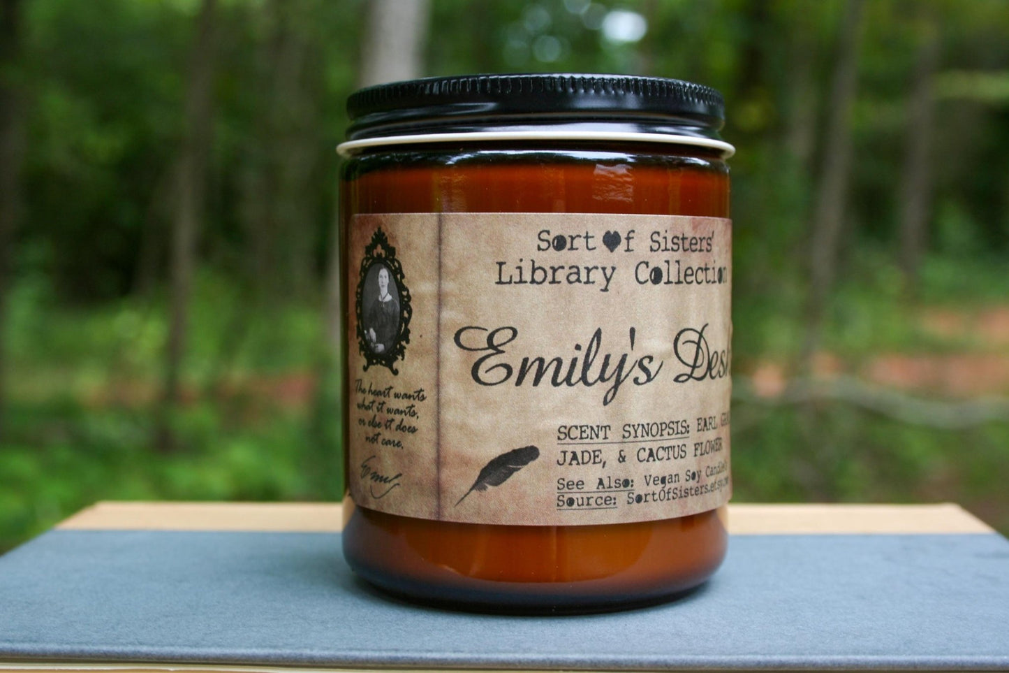 A white soy wax candle in an amber jar with a rectangular label against a white background. The label has like an aged paper look with a binding crease on the left side creating a column. The column has a black and white portrait of Dickinson in a black frame. Under the portrait is a quote and Dickinson's signature. In the larger right column text reads "Sort of Sisters" logo at the top, then "Emily's Desk", then "Earl Grey, Jade, & Cactus Flower", and below that "vegan soy candles, sortofsisters.etsy.com"