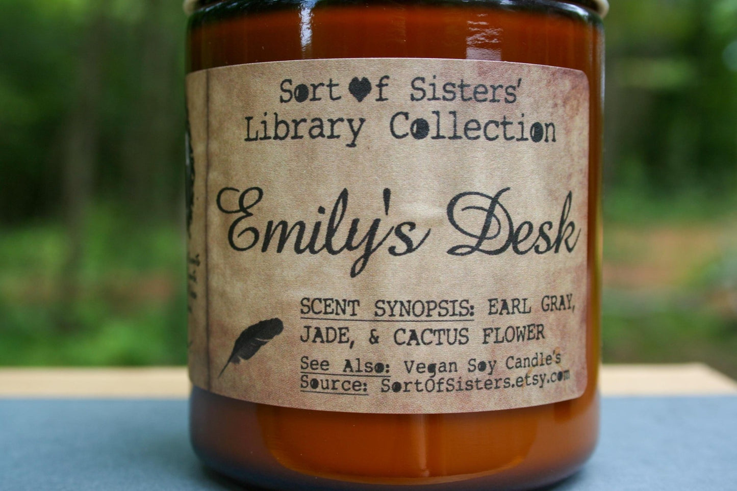 A white soy wax candle in an amber jar with a rectangular label against a white background. The label has like an aged paper look with a binding crease on the left side creating a column. The column has a black and white portrait of Dickinson in a black frame. Under the portrait is a quote and Dickinson's signature. In the larger right column text reads "Sort of Sisters" logo at the top, then "Emily's Desk", then "Earl Grey, Jade, & Cactus Flower", and below that "vegan soy candles, sortofsisters.etsy.com"