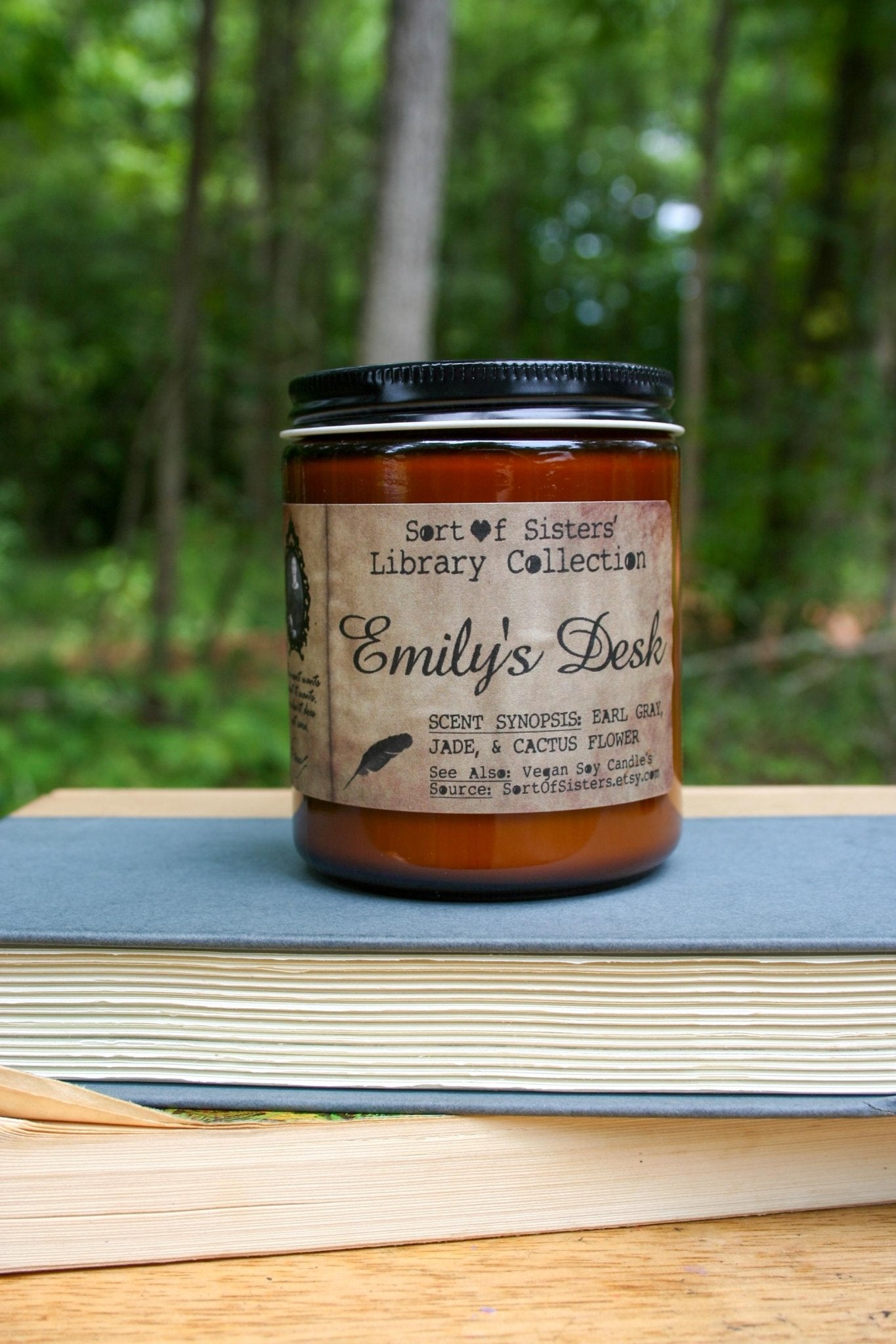 A white soy wax candle in an amber jar with a rectangular label against a white background. The label has like an aged paper look with a binding crease on the left side creating a column. The column has a black and white portrait of Dickinson in a black frame. Under the portrait is a quote and Dickinson's signature. In the larger right column text reads "Sort of Sisters" logo at the top, then "Emily's Desk", then "Earl Grey, Jade, & Cactus Flower", and below that "vegan soy candles, sortofsisters.etsy.com"