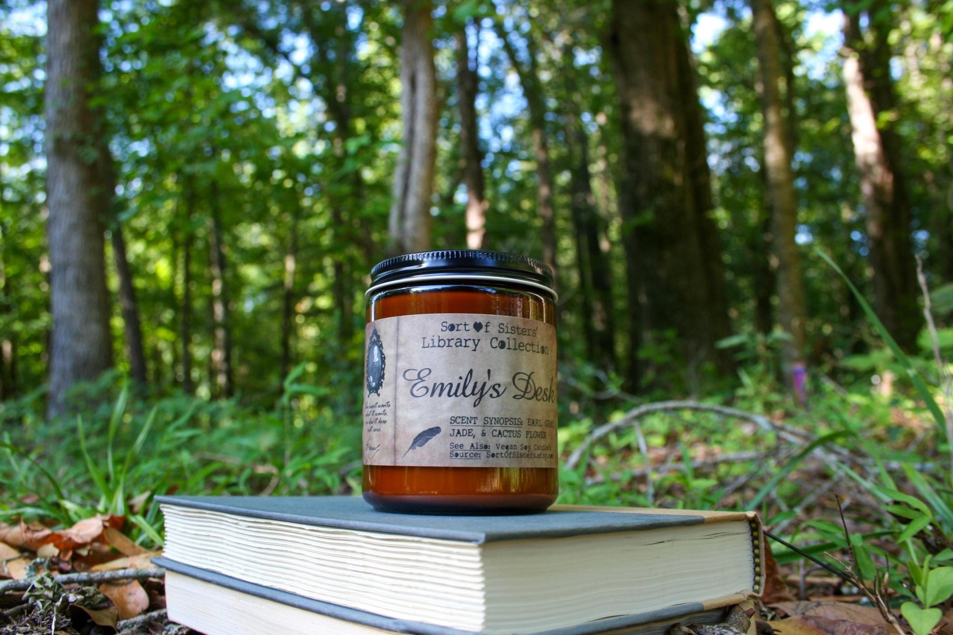 A white soy wax candle in an amber jar with a rectangular label against a white background. The label has like an aged paper look with a binding crease on the left side creating a column. The column has a black and white portrait of Dickinson in a black frame. Under the portrait is a quote and Dickinson's signature. In the larger right column text reads "Sort of Sisters" logo at the top, then "Emily's Desk", then "Earl Grey, Jade, & Cactus Flower", and below that "vegan soy candles, sortofsisters.etsy.com"