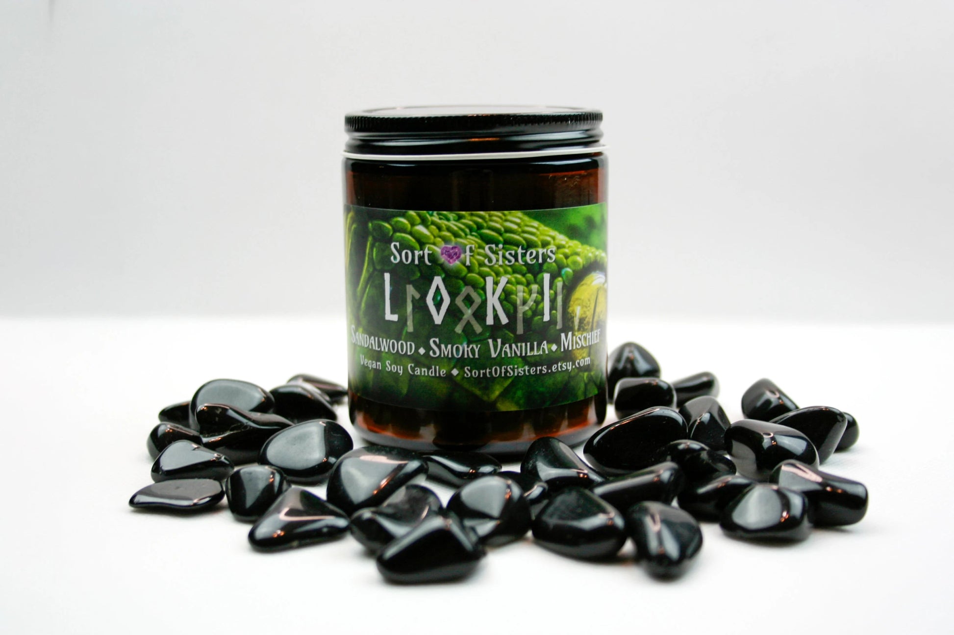 A green soy wax candle in an amber jar with a rectangular label. The label has a green snake on it with yellow eyes. White text on the label reads "Sort of Sisters" logo, below that reads "Loki" (in English and Ruins), below that reads  "sandalwood, smoky vanilla, and mischief", and finally below that reads "vegan soy candle, sortofsisters.etsy.com". The candle is against a white background and surrounded by obsidian stones.