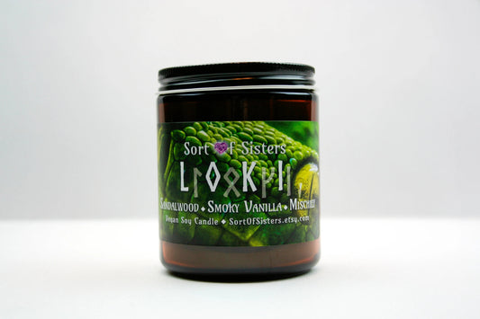 A green soy wax candle in an amber jar with a rectangular label against a white background. The label has a green snake on it with yellow eyes. White text on the label reads "Sort of Sisters" logo, below that reads "Loki" (in English and Ruins), below that reads  "sandalwood, smoky vanilla, and mischief", and finally below that reads "vegan soy candle, sortofsisters.etsy.com".