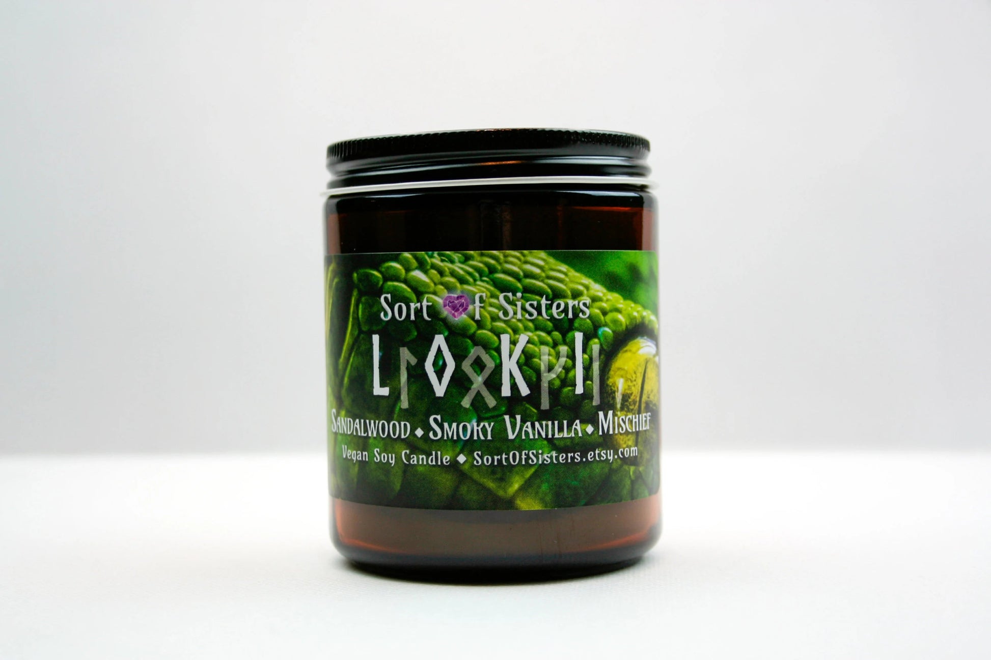 A green soy wax candle in an amber jar with a rectangular label against a white background. The label has a green snake on it with yellow eyes. White text on the label reads "Sort of Sisters" logo, below that reads "Loki" (in English and Ruins), below that reads  "sandalwood, smoky vanilla, and mischief", and finally below that reads "vegan soy candle, sortofsisters.etsy.com".